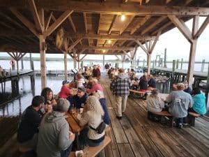 People sitting at tables on the dock at the Bayshore Center at Bivalve during a 2nd Friday community event, enjoying food, drinks, and waterfront views in Port Norris, New Jersey.