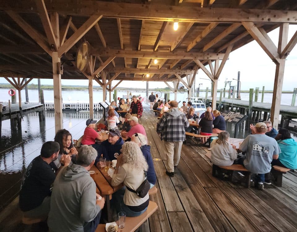 People sitting at tables on the dock at the Bayshore Center at Bivalve during a 2nd Friday community event, enjoying food, drinks, and waterfront views in Port Norris, New Jersey.
